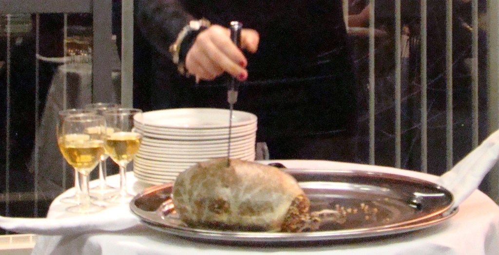 Picture of haggis on a serving plate being pierced by a dagger. Drams of whiskey are in four small glasses nearby.
